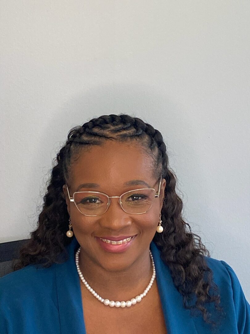 A woman with glasses and braids sitting in front of a wall.